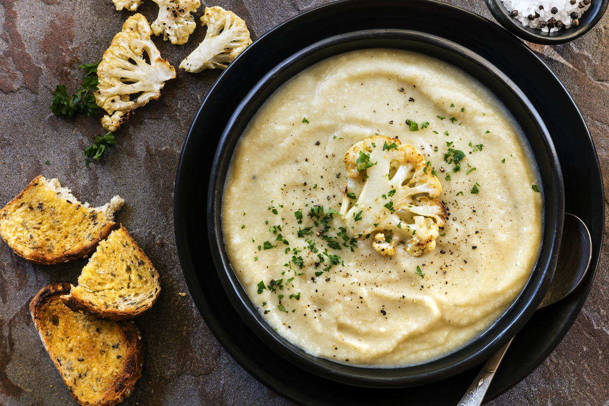 Eine Schüssel Karfiolcremesuppe, garniert mit gerösteten Karfiolröschen und frischen Kräutern, daneben geröstete Brotscheiben.