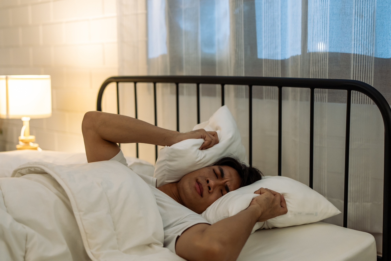 Mann liegt im Bett und hält ein Kissen über seinen Kopf.