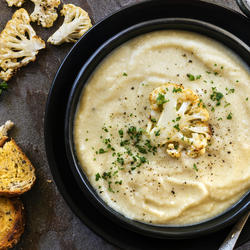 Eine Schüssel Karfiolcremesuppe, garniert mit gerösteten Karfiolröschen und frischen Kräutern, daneben geröstete Brotscheiben.