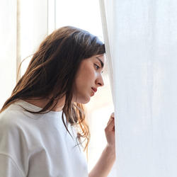Frau schaut traurig aus einem Fenster hinter einem weißen Vorhang