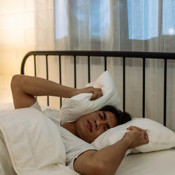 Mann liegt im Bett und hält ein Kissen über seinen Kopf.